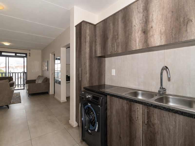 Modern kitchen area featuring wood-look cabinetry, granite countertops, a double bowl sink, and tiled flooring, seamlessly flowing into the adjacent living space.