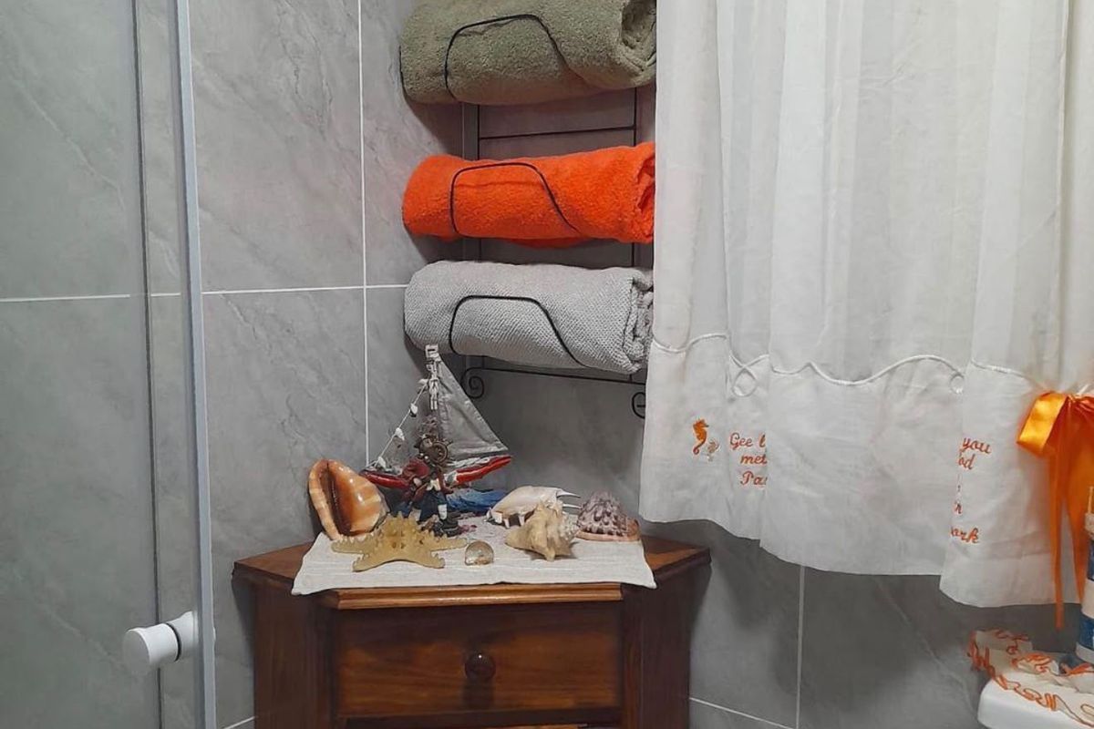 A bathroom corner featuring grey wall tiles, a shower enclosure, and a toilet, with a wooden corner cabinet providing storage.