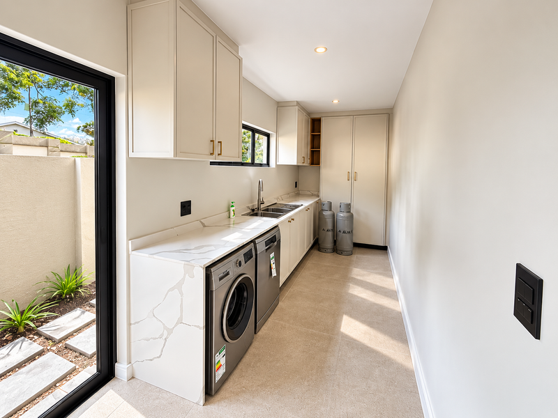 A modern, well-lit scullery featuring sleek built-in cabinetry, stone countertops, a double sink, and direct access to an outdoor area through a sliding door.