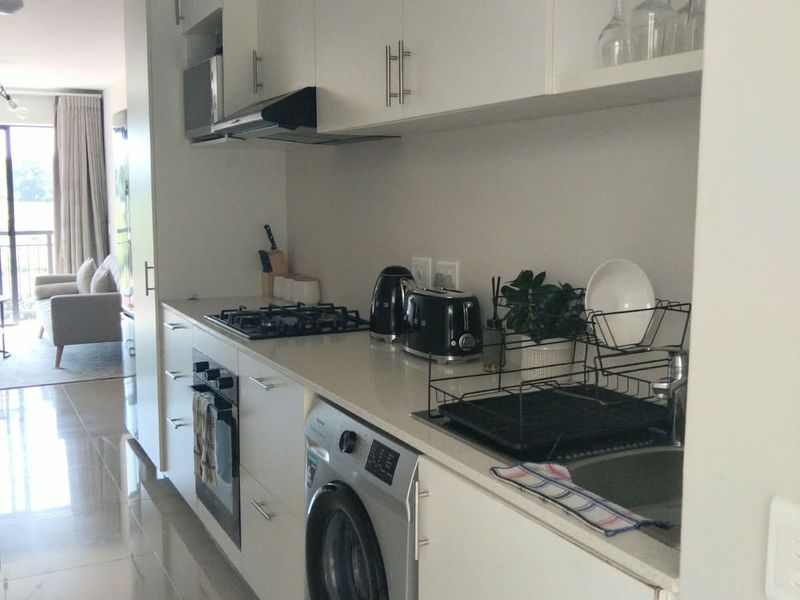 A modern, compact galley-style kitchen featuring white cabinetry, a gas hob, built-in oven, and integrated laundry appliance, finished with sleek tiled flooring.