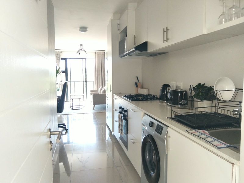 Modern galley-style kitchen featuring white cabinetry, stone countertops, integrated appliances, and sleek tiled flooring, leading into an open-plan living area.