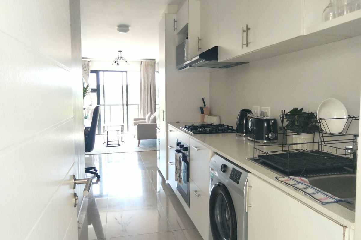 Modern galley-style kitchen featuring white cabinetry, stone countertops, integrated appliances, and sleek tiled flooring, leading into an open-plan living area.