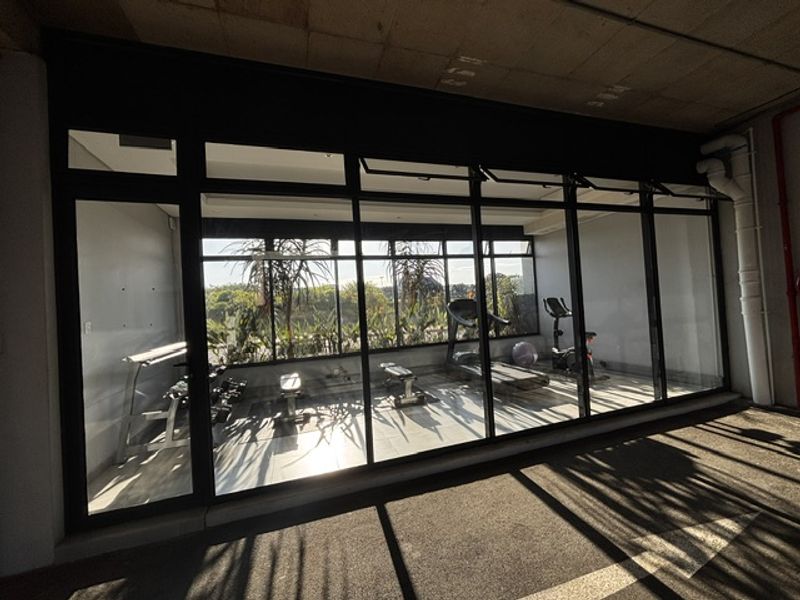 A modern gym facility featuring floor-to-ceiling glass windows, tiled flooring, and an exposed concrete ceiling, providing a bright and functional workout space.