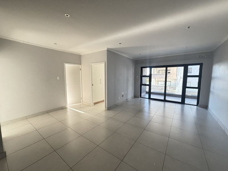 A spacious, empty living area featuring neutral tiled flooring, recessed lighting, and large sliding glass doors leading to an outdoor balcony.
