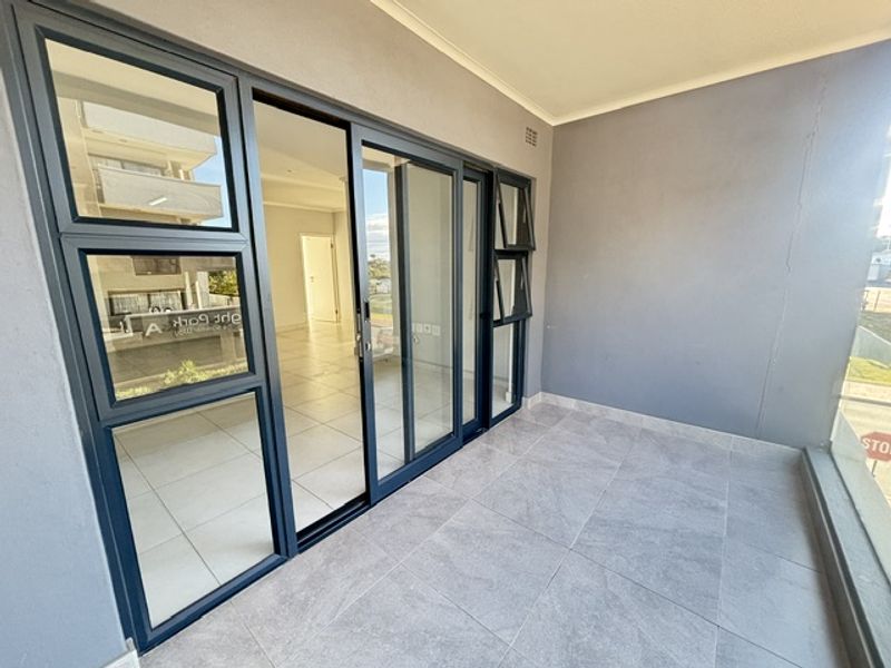 A modern, covered balcony featuring large sliding glass doors, neutral tiled flooring, and clean grey walls, offering a seamless transition to the interior living space.