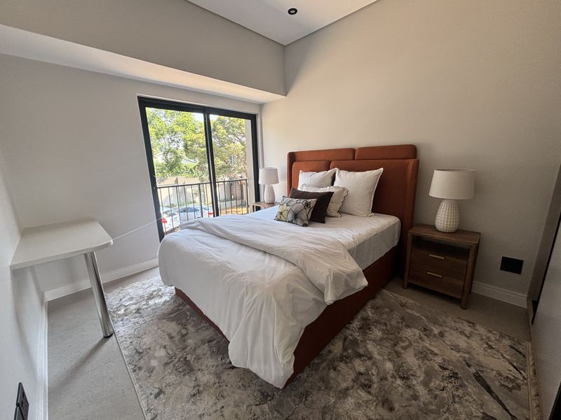 A modern bedroom featuring neutral walls, recessed lighting, a built-in desk, and a sliding glass door leading to a private balcony.