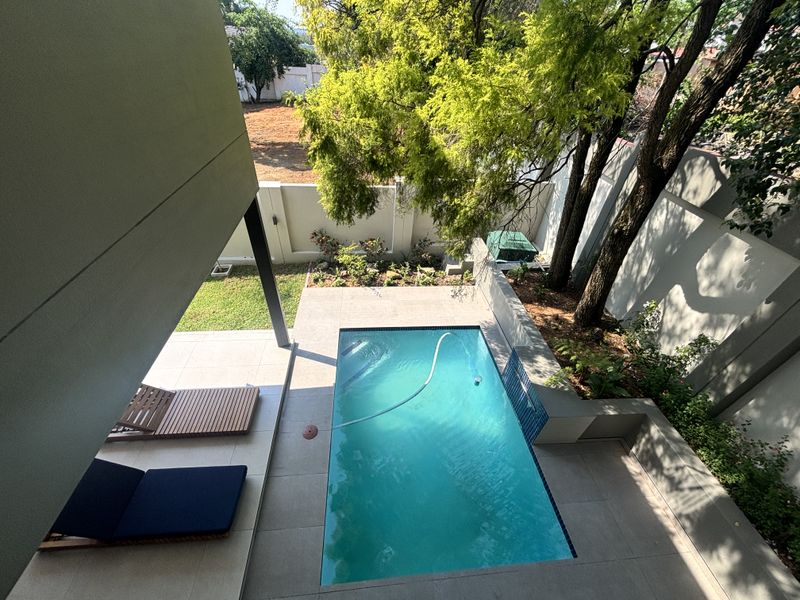 An elevated view of a private swimming pool area featuring a tiled patio, boundary walls, and established trees in a landscaped garden setting.