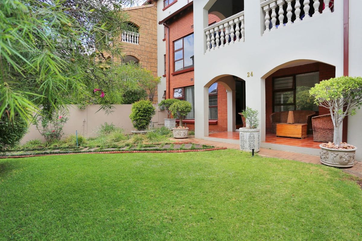 A well-maintained rear exterior view featuring a lush green lawn, a covered patio with tiled flooring, and a classic brick and plaster facade.