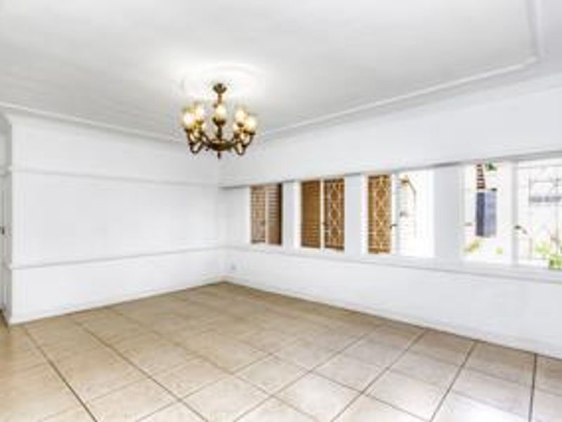 A bright, empty living room featuring tiled flooring, a decorative ceiling rose with a chandelier, and large windows with picture rail detailing.
