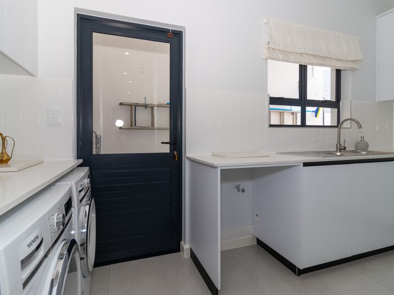 A clean and functional laundry room featuring white cabinetry, a stainless steel sink, tiled splashbacks, and direct access to the exterior.