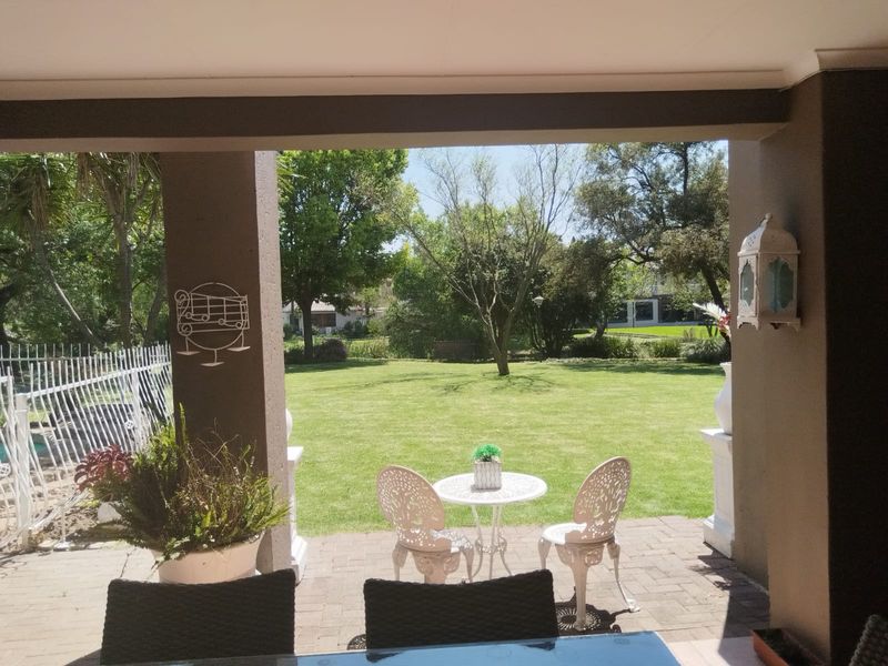 A covered patio area featuring paved flooring and structural pillars, overlooking a spacious, well-maintained garden.