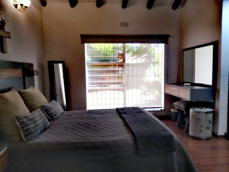 A cozy bedroom featuring exposed timber ceiling beams, laminate flooring, and a wall-mounted vanity unit, with natural light entering through a security-barred window.