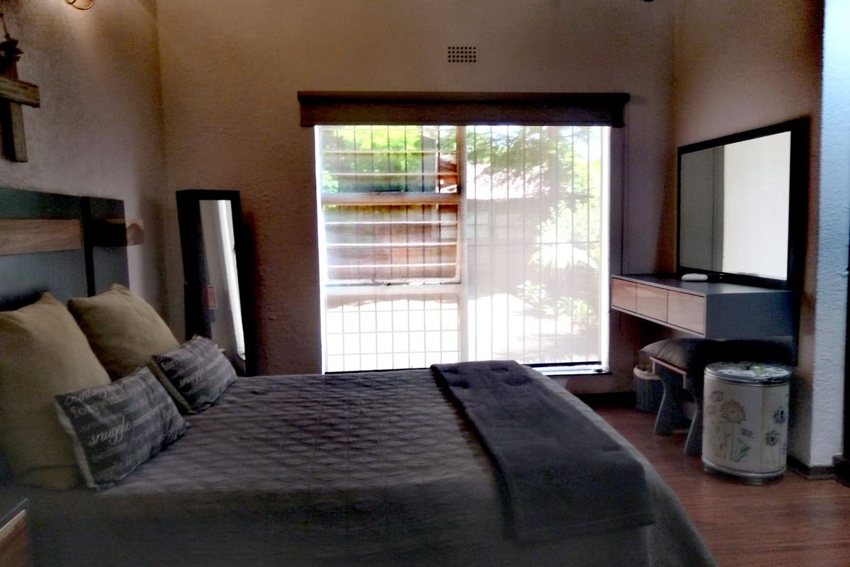 A cozy bedroom featuring exposed timber ceiling beams, laminate flooring, and a wall-mounted vanity unit, with natural light entering through a security-barred window.