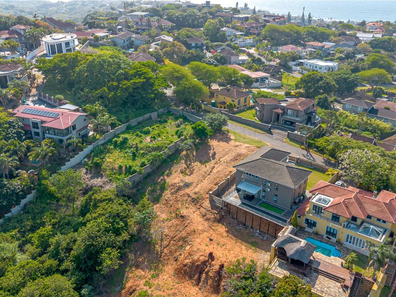 Aerial view of a vacant residential plot situated in a developed suburban neighborhood, featuring sloped terrain and established boundary walls.