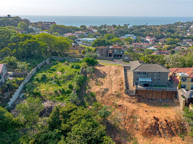 Aerial view of a vacant residential plot featuring sloping terrain, boundary walls, and surrounding established neighborhood with distant ocean views.