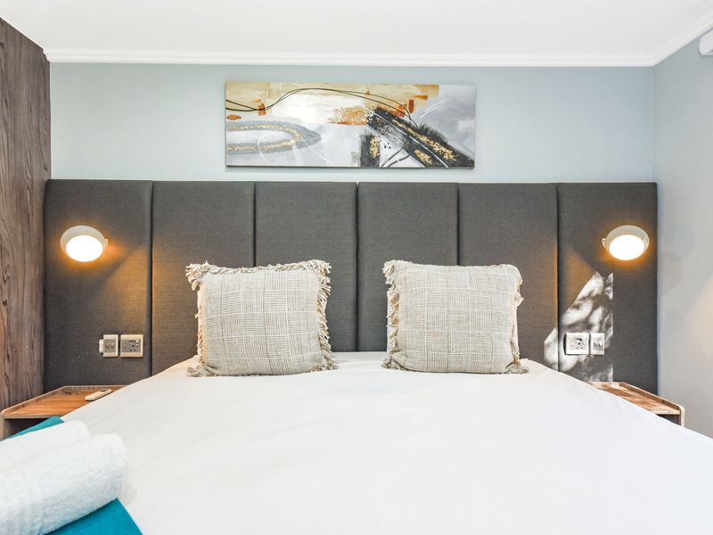 A modern bedroom featuring a large upholstered headboard, integrated wall-mounted reading lights, and built-in bedside shelving with a clean, contemporary finish.