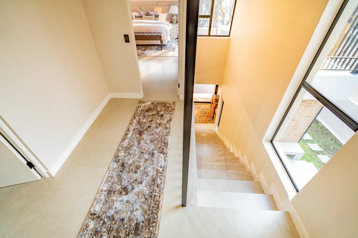 A bright, modern hallway featuring tiled flooring, a staircase with a metal handrail, and large windows providing natural light and views of the exterior.