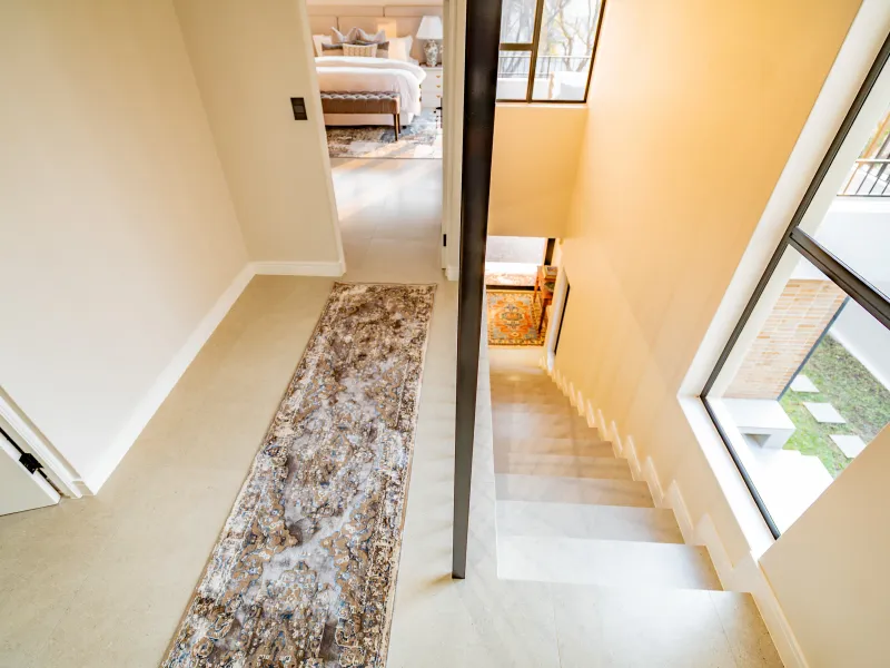 A bright, modern hallway featuring tiled flooring, a staircase with a metal handrail, and large windows providing natural light and views of the exterior.