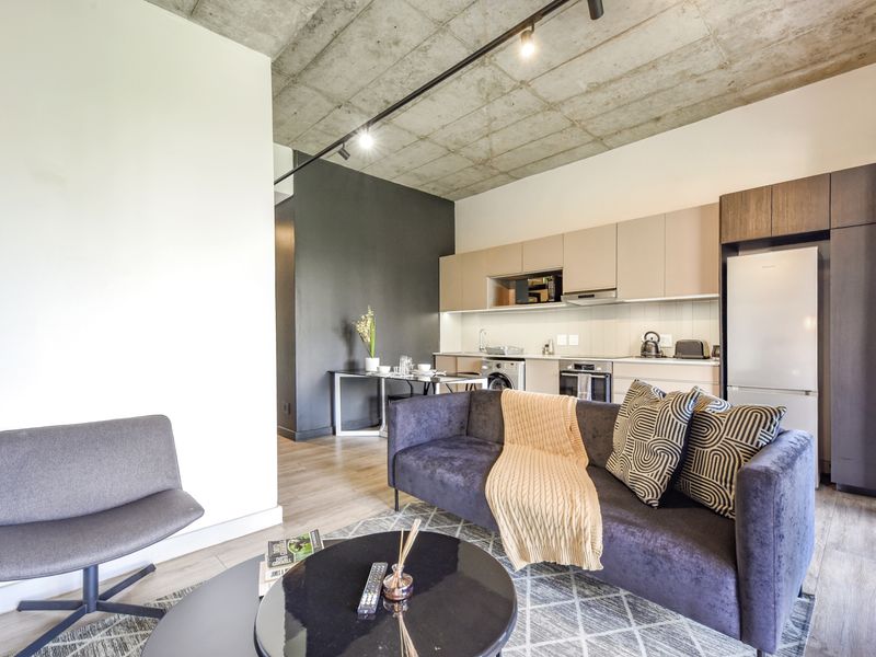 Modern open-plan living area featuring an exposed concrete ceiling, track lighting, and a sleek, well-equipped kitchen with integrated appliances.