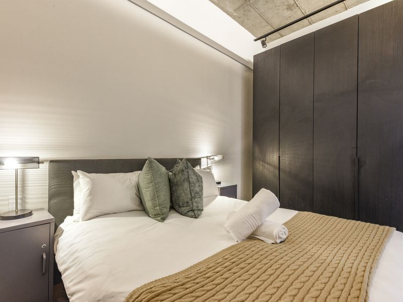 Modern bedroom featuring a sleek dark built-in wardrobe, exposed concrete ceiling with track lighting, and ambient recessed wall lighting.