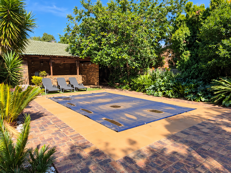 A well-maintained outdoor pool area featuring a covered patio, brick paving, and established garden landscaping.