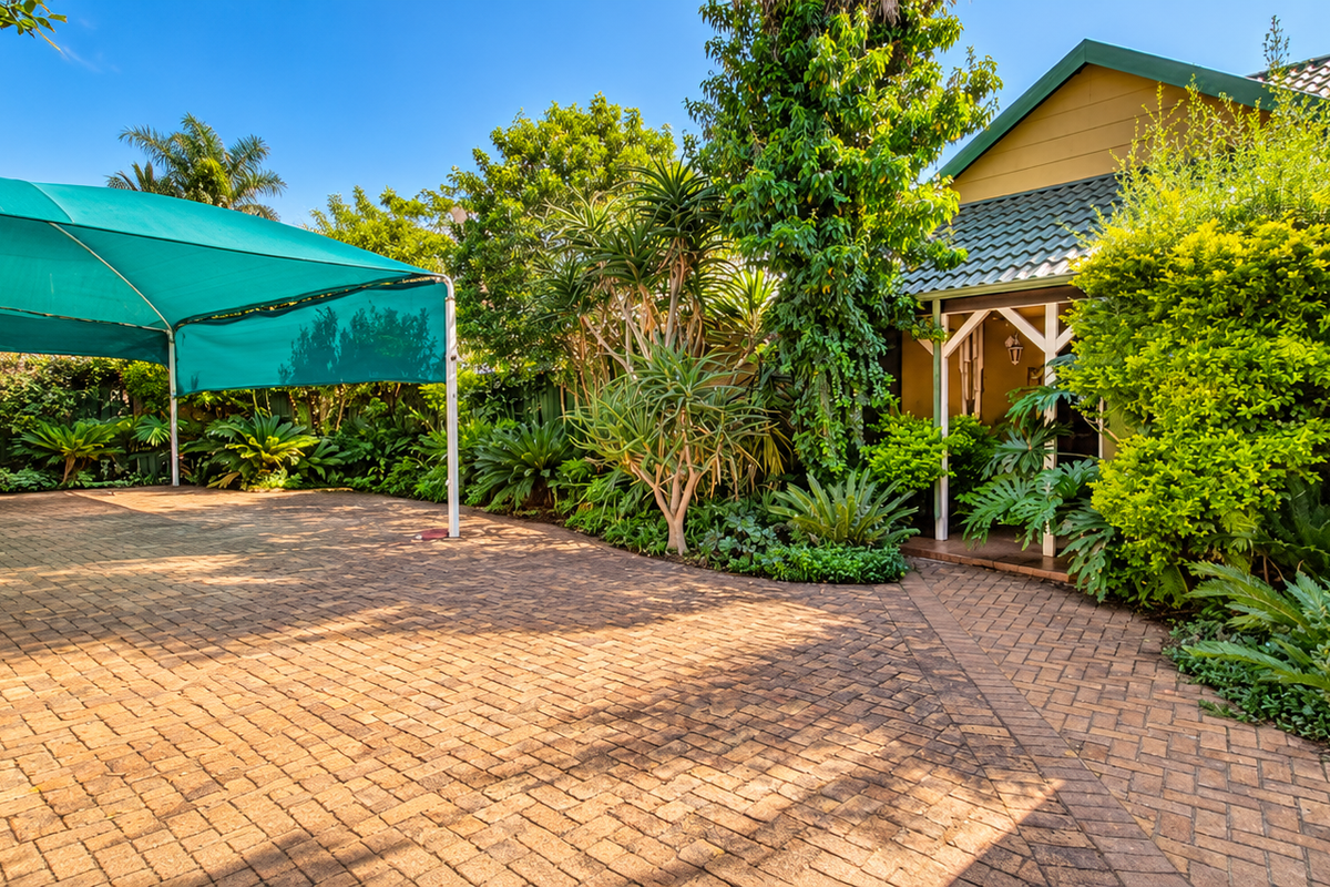 A well-maintained rear exterior area featuring a paved courtyard, a large green shade sail, and established garden landscaping leading to the home entrance.