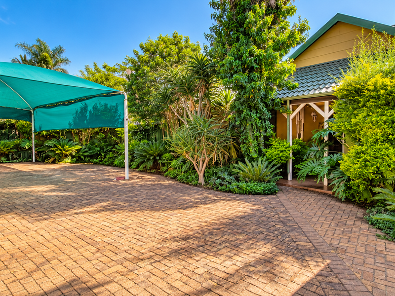A well-maintained rear exterior area featuring a paved courtyard, a large green shade sail, and established garden landscaping leading to the home entrance.