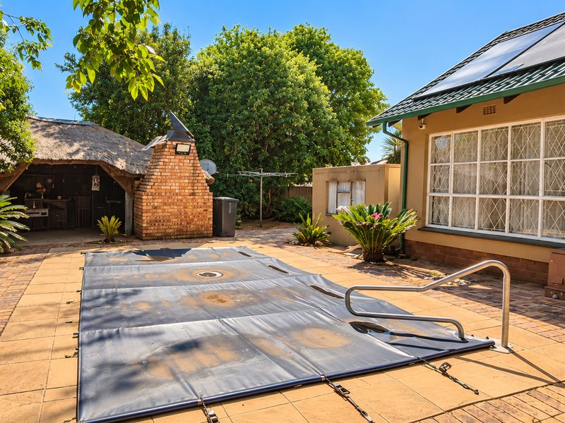 Outdoor pool area featuring a paved patio, a covered swimming pool, a brick braai, and a thatched lapa, set within a landscaped garden.