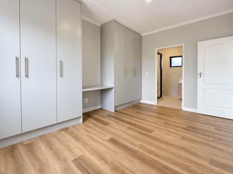 A bright, modern bedroom featuring light-toned laminate flooring, extensive built-in cabinetry with an integrated desk, and direct access to an en-suite bathroom.