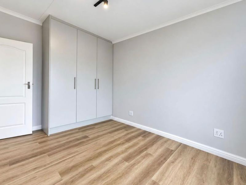 A bright, empty bedroom featuring modern light-toned laminate flooring, neutral grey walls, and built-in cupboards with clean lines.