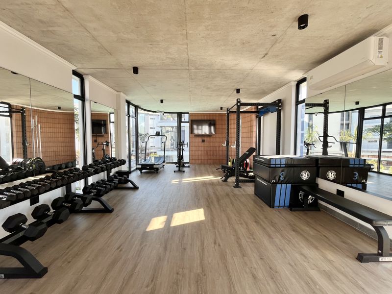 Modern apartment gym featuring an exposed concrete ceiling, wood-look flooring, full-height mirrors, and a stylish terracotta-tiled feature wall.