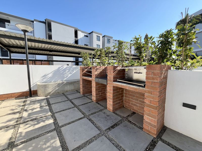Outdoor patio area featuring a built-in brick braai, paved flooring, and a covered structure, ideal for entertaining in a modern apartment complex setting.