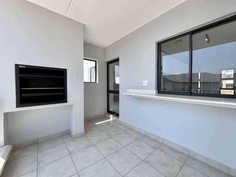 A clean, modern covered balcony featuring tiled flooring, a built-in braai, and a convenient serving shelf, perfect for outdoor entertaining.