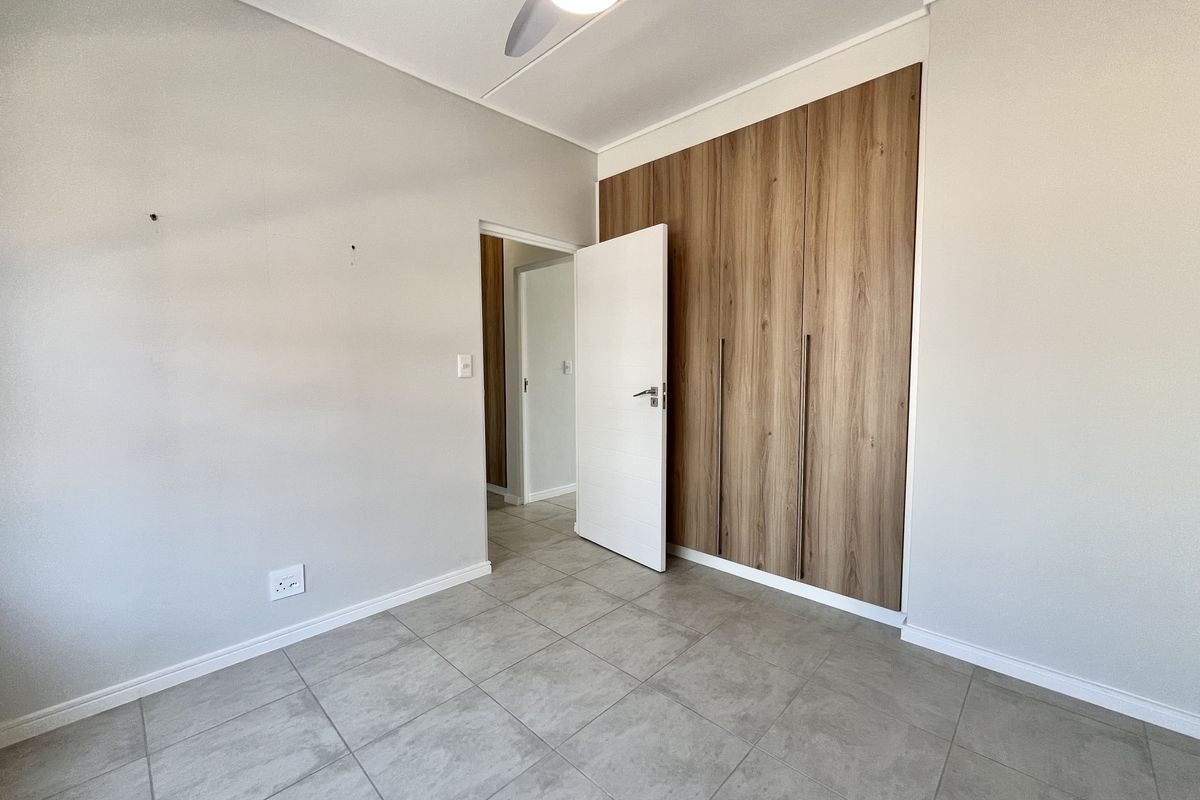 A bright, empty bedroom featuring modern wood-finish built-in cupboards, neutral tiled flooring, and a ceiling fan.