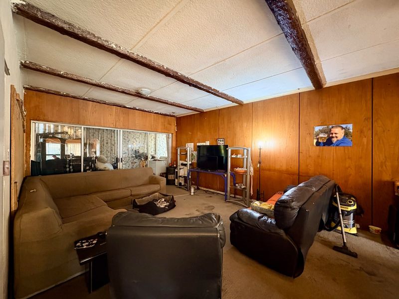 Spacious living room featuring exposed timber ceiling beams, wood-paneled walls, and large sliding glass doors, offering a dated interior ready for renovation.