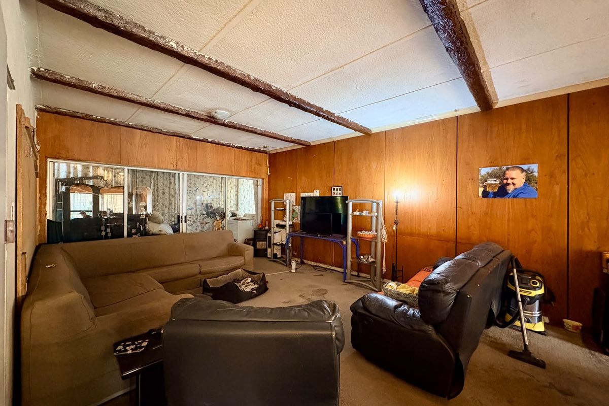 Spacious living room featuring exposed timber ceiling beams, wood-paneled walls, and large sliding glass doors, offering a dated interior ready for renovation.