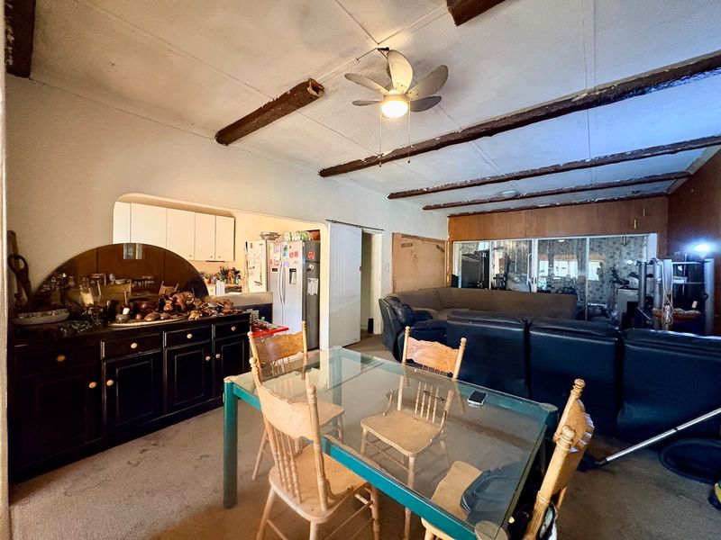 Open plan living and dining area featuring exposed timber ceiling beams, dated finishes, and direct access to the kitchen space.