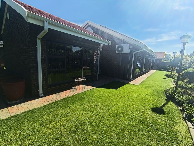 A well-maintained rear exterior view featuring a brick facade, paved walkway, and neatly landscaped artificial lawn with integrated garden lighting.