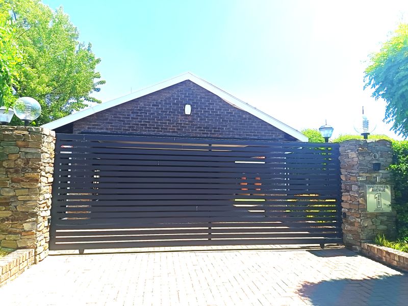 Front entrance view featuring a modern horizontal slat sliding gate, stone pillars, and a brick driveway.