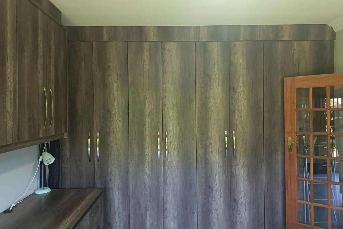 A bedroom featuring extensive built-in cupboards with a wood-grain finish, a matching built-in desk, and a classic timber-framed glass door.