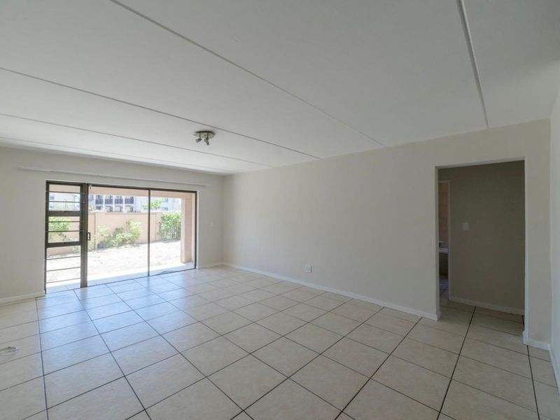 A spacious, empty living room featuring neutral tiled flooring, a sliding glass door leading to the exterior, and a clean, minimalist aesthetic.