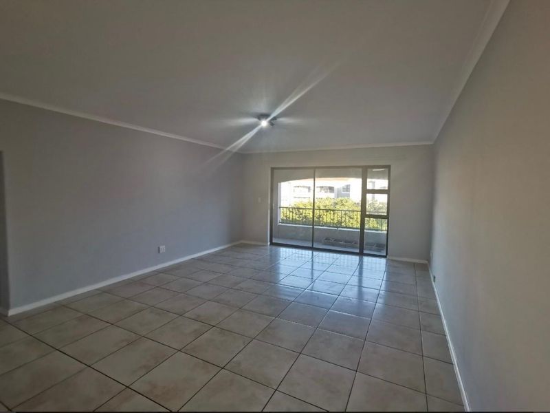 A spacious, empty living room featuring neutral grey walls, ceramic floor tiles, and a large sliding glass door providing access to a balcony.