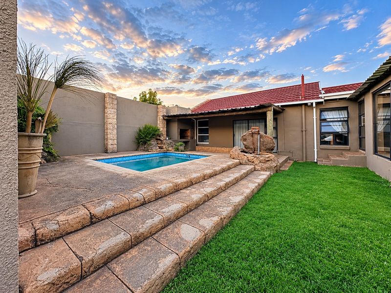 Outdoor entertainment area featuring a swimming pool, built-in braai, and stone-clad steps leading to a well-maintained garden and rear building elevation.