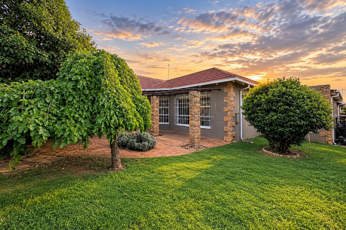 A well-maintained rear exterior view featuring a brick-paved patio, stone-clad pillars, and a lush garden under a sunset sky.