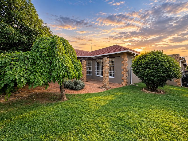 A well-maintained rear exterior view featuring a brick-paved patio, stone-clad pillars, and a lush garden under a sunset sky.