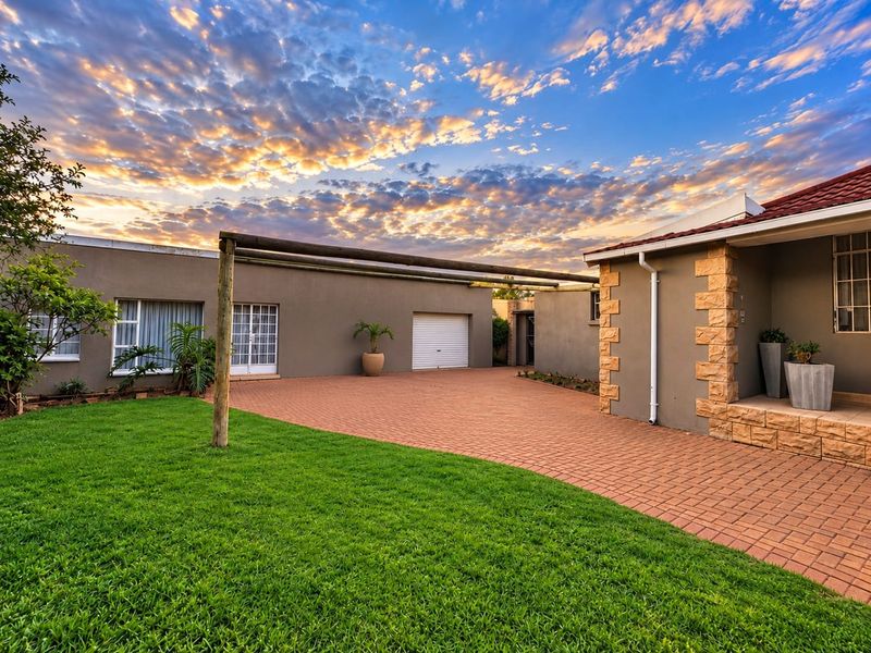 Front exterior view featuring a paved driveway, stone-clad accents, and a timber pergola, set against a well-maintained lawn and sunset sky.