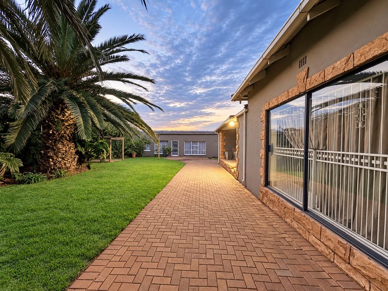 A well-maintained rear exterior view featuring a paved walkway, manicured lawn, and stone-clad facade with secure sliding glass doors at sunset.