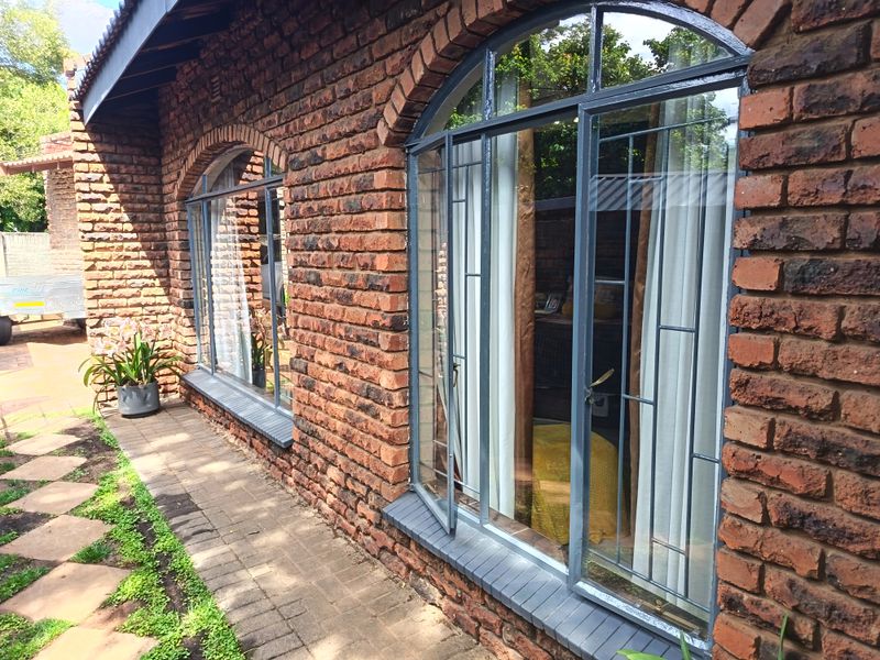 A well-maintained rear exterior view featuring exposed brickwork, arched steel-framed windows with security bars, and a paved garden walkway.