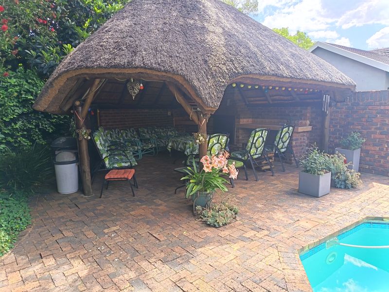A covered outdoor patio area featuring a traditional thatch roof, brick paving, and a built-in braai, situated adjacent to the swimming pool.