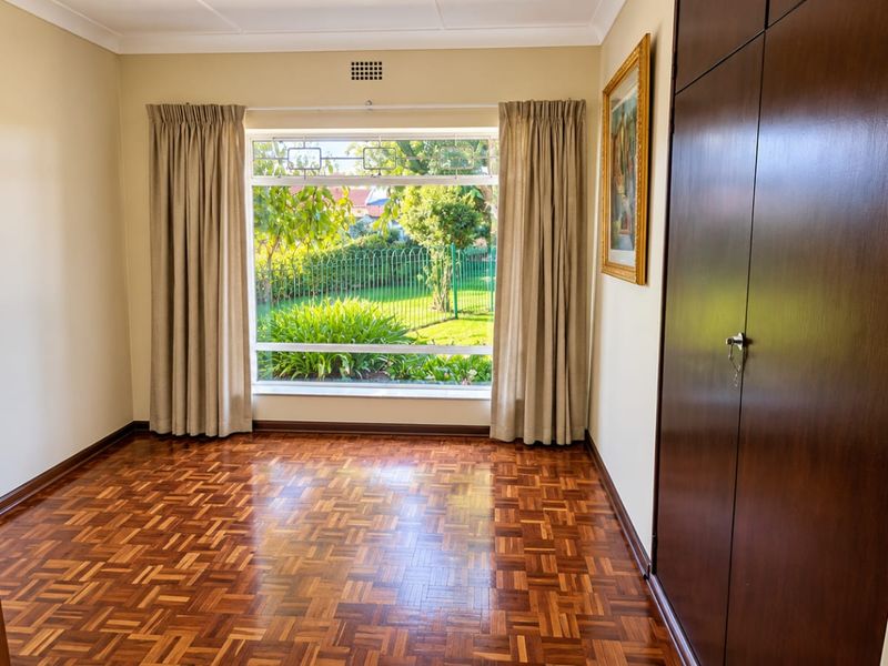 A bright, empty bedroom featuring classic parquet flooring, built-in wooden cupboards, and a large window overlooking the garden.